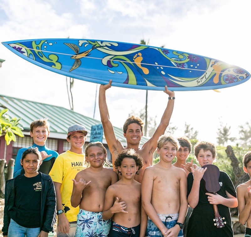 Kai Lenny holding a custom surfboard aloft that will be auctioned to benefit the Paia Youth and Cultural Center